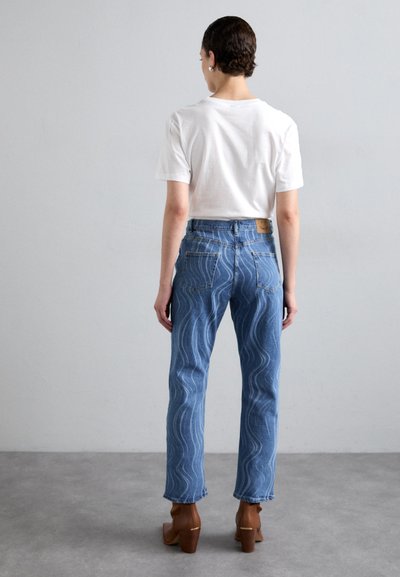 Lyseblå jeans med en bølgende hvit søm, høyt liv og et lærmerke på baklivet. Kombinert med en hvit t-skjorte.