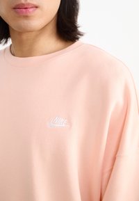 Ljusrosa sweatshirt i mjukt material med en vit Nike-logotyp på bröstet. Rund halsringning och avslappnad passform.