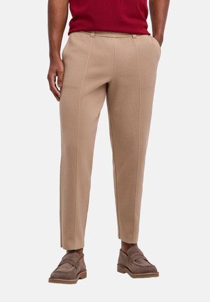 Mann trägt beige maßgeschneiderte Hose und braune Wildleder-Loafer mit rotem Hemd, steht mit Händen in den Taschen vor einfarbigem Hintergrund.
