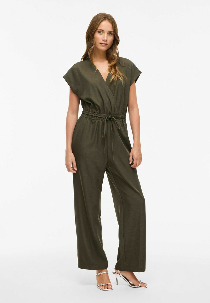 Mujer vestida con un mono sin mangas de color verde oliva, con cintura ajustable y sandalias de tacón alto plateadas, de pie contra un fondo blanco.
