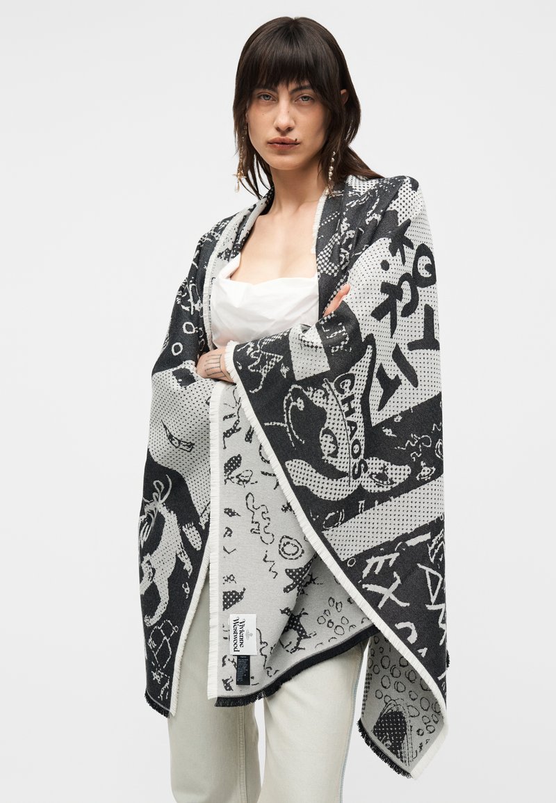 Femme aux cheveux foncés porte un châle noir et blanc à motifs sur un haut blanc et un pantalon clair, debout devant un fond uni.