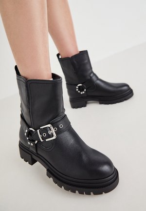 Schwarze Leder-Stiefeletten mit klobigen Sohlen und silbernen Schnallenriemen, getragen an nackten Beinen vor einem schlichten hellen Hintergrund.