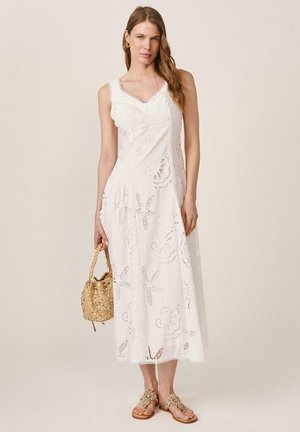 Mujer con vestido midi blanco sin mangas con recortes florales, sosteniendo un bolso beige con tachuelas y usando sandalias con joyas, de pie frente a un fondo liso.