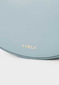 Svijetloplava okrugla kožna torba s glatkom teksturom, s zlatno utisnutim "FURLA" logom. Vidljivo šivanje uz rub.