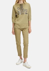 Olivově zelená mikina s výrazným textovým designem, doplněná o světlé béžové zúžené kalhoty s knoflíkovými detaily a bílé tenisky.