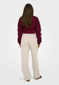 Burgunderfarbener, übergroßer Sweatshirt mit weißem Grafikdruck auf dem Ärmel, kombiniert mit beigen, weit geschnittenen Jogginghosen mit sichtbarem Gummibund.