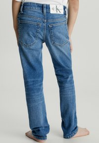 Vaqueros de mezclilla azul con corte recto, que presentan bolsillos traseros y una etiqueta de Calvin Klein en la cintura. La textura es suave y ligeramente desgastada.