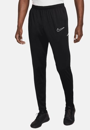 Mężczyzna ubrany w czarne spodnie dresowe Nike z białym logo, czarne buty sportowe i białą koszulę, stoi na jednolitym białym tle.