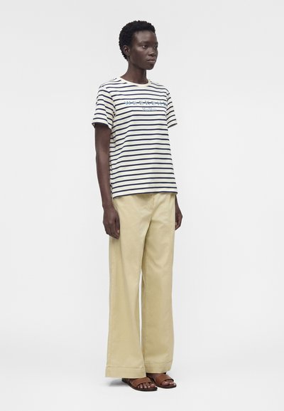 Άτομο που φοράει λευκό και μπλε ριγέ μπλουζάκι με κείμενο, μπεζ φαρδιά παντελόνια και καφέ σανδάλια, στέκεται μπροστά σε μια μονόχρωμη επιφάνεια.