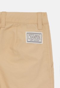 Bež pamučne šortse s pravokutnom tkanom etiketom na kojoj stoji "Champion Dry Goods" u tamnoplavoj i bijeloj boji. Uključuje petlje za remen i bočne džepove.