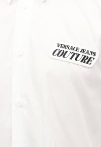 Άσπρο πουκάμισο με κουμπιά, με μαύρο και λευκό κέντημα που γράφει "VERSACE JEANS COUTURE" στην περιοχή της τσέπης στο στήθος.