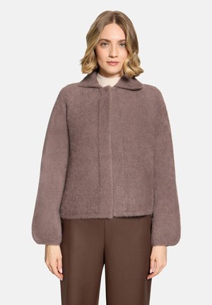 Vrouw met schouderlang blond haar die een mauve zachte jas met lange mouwen en een bruine broek draagt tegen een eenvoudige lichte achtergrond.