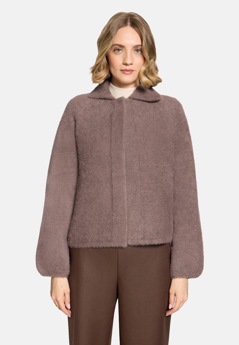 Femme aux cheveux blonds mi-longs portant une veste duveteuse mauve à manches longues et un pantalon marron sur un fond clair uni.
