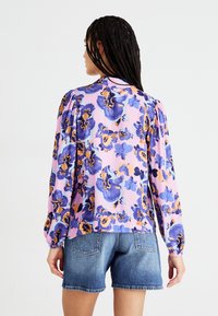 Blusa a maniche lunghe in tessuto rosa con un grande motivo floreale in blu e arancione. Presenta polsini elasticizzati e una vestibilità comoda.