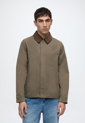 Ung mand iført en brun jakke med en mørkebrun krave og blå jeans, stående mod en ensfarvet hvid baggrund.