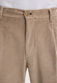 Zbliżenie na beżowe spodnie sztruksowe z zapinanym na guzik stanem i widocznymi szlufkami na pasek, noszone z białym topem.