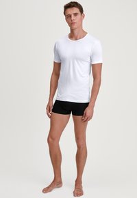 White short-sleeve t-shirt made of soft fabric, fitted design. Paired with black boxer briefs, standing barefoot on a neutral background.