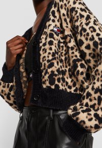 Cardigan en imprimé léopard en tissu tricoté doux, avec des poignets et un ourlet côtelés noirs, des boutons sur le devant et une petite étiquette logo sur la poitrine.