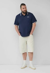 Marineblaue Poloshirt mit weißen Streifen am Kragen und an den Ärmeln, kombiniert mit cremefarbenen Shorts und weißen Turnschuhen. Einfaches und lässiges Design.