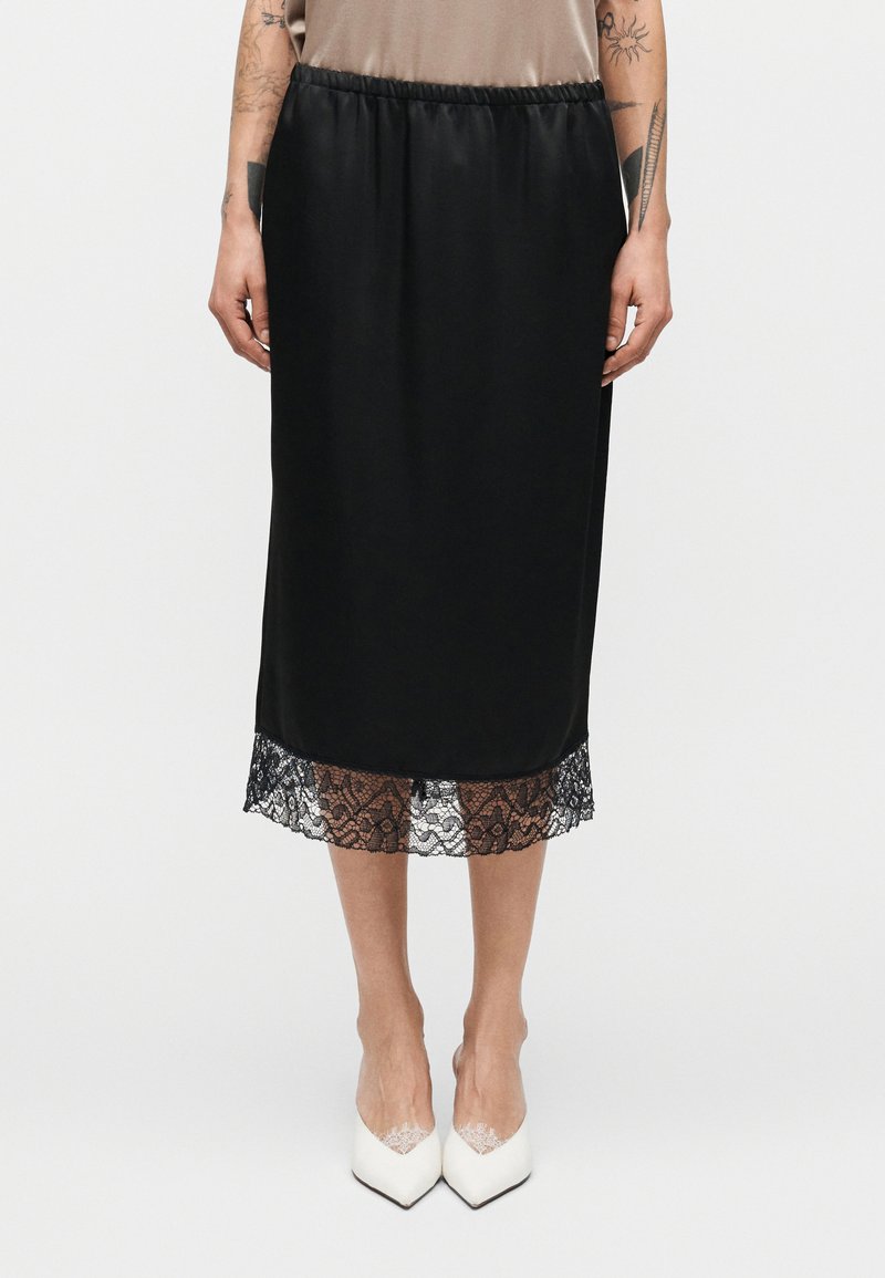 Μαύρη μίντι φούστα με λεία σατινέ υφή, διακοσμημένη με δαντέλα περιοχής με κυματιστό σχέδιο στο τελείωμα. Ελαστική μέση για άνεση. Λευκά μυτερά παπούτσια.