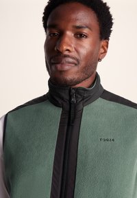 Green fleece vest with black accents, high collar, front zipper, and logo on the chest. Soft texture and contrasting colors.