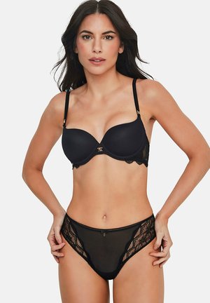 Femme aux longs cheveux noirs portant un soutien-gorge en dentelle noire et une culotte taille haute assortie, debout les mains sur les hanches devant un arrière-plan uni.