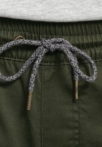 Los pantalones cortos de algodón verde presentan un cordón con un patrón de espiga gris y un detalle de herrajes dorados. Tela texturada y costuras visibles.