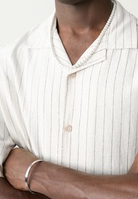 Chemise à manches courtes blanche rayée avec col à revers, dotée d'une fermeture à boutons sur le devant. Accentuée par un collier de perles et un bracelet en métal.