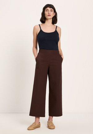 Woman standing with hands in pockets wearing a black tank top, wide-leg brown pants, and woven slip-on shoes on a light background.