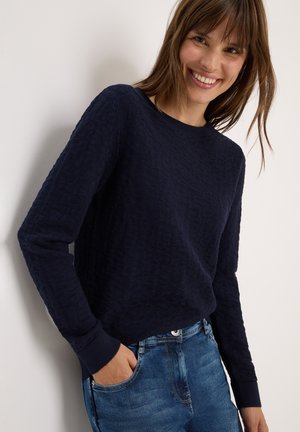 Mørkeblå struktureret sweater med rund hals og lange ærmer, parret med lyse denimjeans. Sweaterens har subtile mønstre og ribbede manchetter.