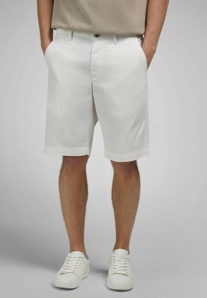 Man wearing beige knit shirt, white knee-length shorts with pockets, and white sneakers, standing with hands in pockets against gray background.