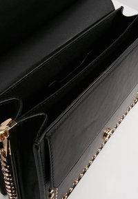 Borsa a tracolla in pelle nera con dettagli in oro, scomparti interni e borchie decorative lungo il bordo anteriore. Texture liscia e design strutturato.