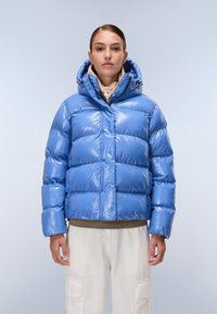 Skinnende blå pufferjakke med en aftagelig hætte, der har en boxy skæring, quiltet tekstur og lynlås foran. Bæret over en beige top.