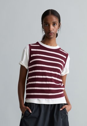 Femme aux cheveux foncés attachés, portant un pull sans manches rayé bordeaux et blanc par-dessus un t-shirt blanc et un pantalon noir, les mains dans les poches.