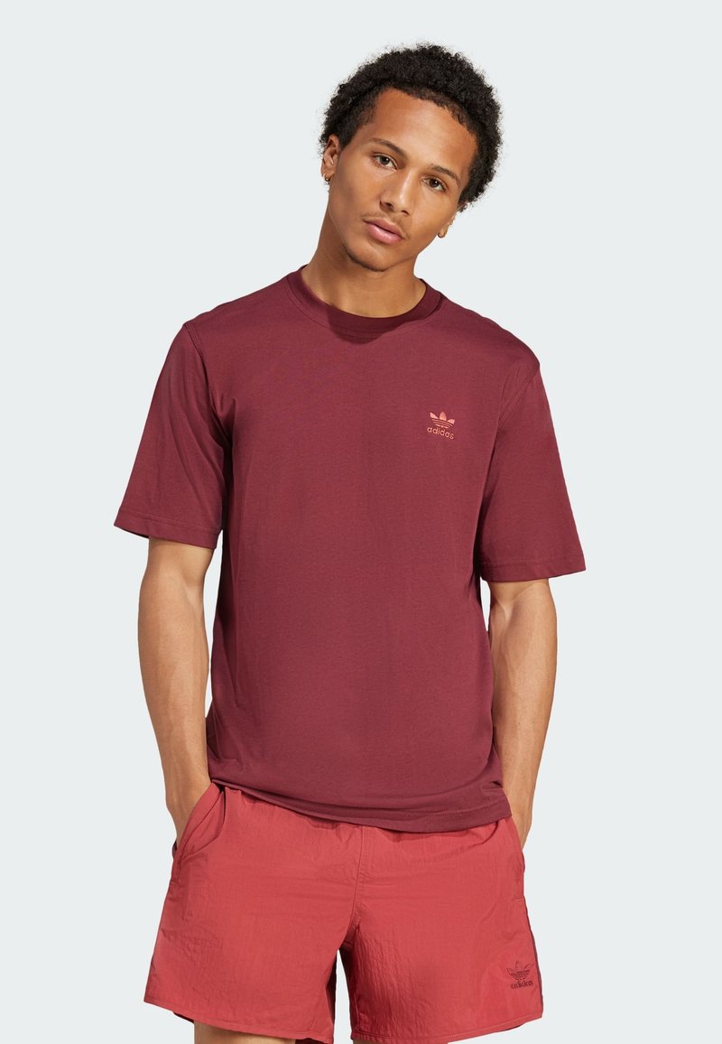 Burgundska bombažna majica s kratkimi rokavi in majhnim svetlo rožnatim logotipom Adidas na levem prsnem delu, v paru s svetlo rdečimi kratkimi hlačami.