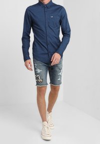 Námořnická košile na knoflíky s kapsou; džínové šortky s roztrženými detaily a grafickými nášivkami; krémové tenisky. Jednoduchý design a ležérní střih.