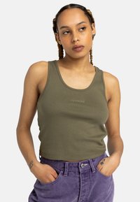 Olivengrön ribbad linne med en figurnära design. Har en rundad halsringning och subtil logotypdetaljering. Står i kombination med högmidjade lila jeans.