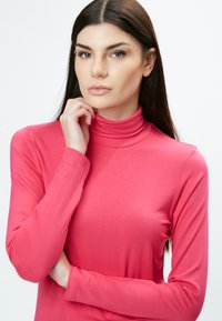 Langärmliges Rollkragenoberteil in leuchtendem Pink, aus glattem Stoff mit schmaler Silhouette. Verfügt über einen hohen, gefalteten Kragen und eng anliegende Ärmel.