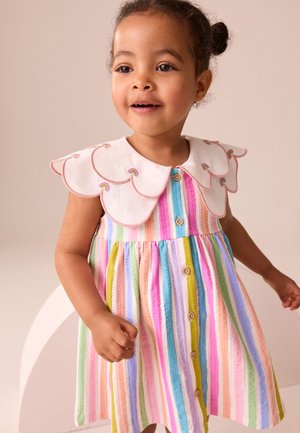 Jeune fille souriante portant une robe colorée à rayures verticales avec des boutons en bois et un grand col blanc festonné orné d'une broderie arc-en-ciel.