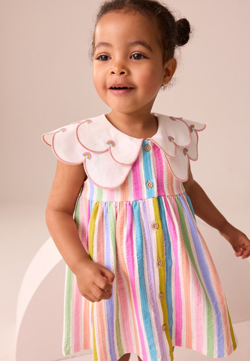 Jeune fille souriante portant une robe colorée à rayures verticales avec des boutons en bois et un grand col blanc festonné orné d'une broderie arc-en-ciel.