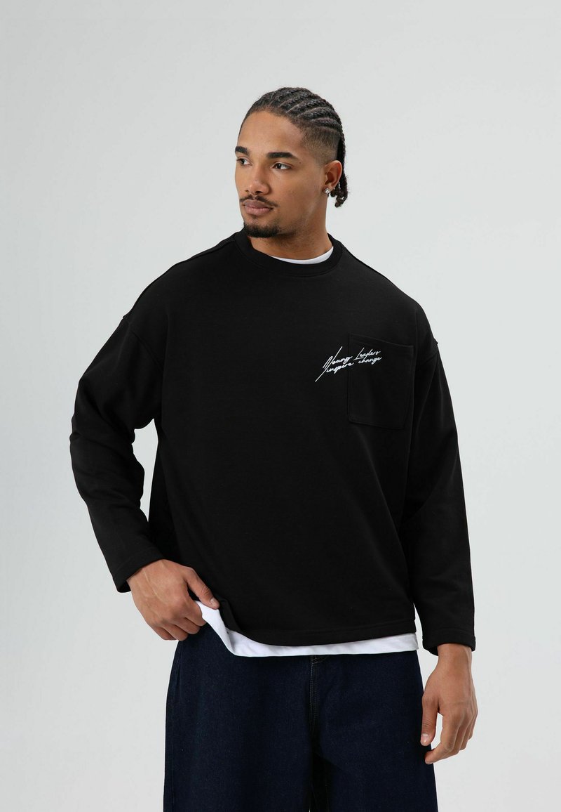 Zwart shirt met lange mouwen, een ronde hals en een borstzak aan de voorkant. Voorzien van witte geborduurde tekst op het zakgebied.