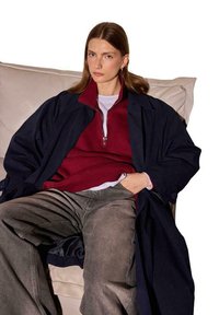 Jūrinis paltas virš burgundinės spalvos zip-up megztinio, derinamas su baltu marškinėliu ir pilkomis džinsomis. Lygi tekstūra ir laisvas kirpimas.