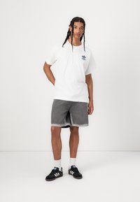 Hvit bomull t-skjorte med blå logo, grå denimshorts og svarte sneakers med hvite detaljer. Modellen står med den ene hånden på hoften.