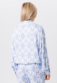 Vrouw met lang blond haar, gekleed in een losse witte blouse met lichtblauwe patronen en bijpassende broek, gezien vanaf de achterkant.