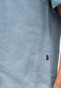 Ανοιχτό μπλε πουκάμισο από λινό με κοντά μανίκια και μια μικρή μαύρη ετικέτα με την ένδειξη «BOSS» κοντά στην πλαϊνή ραφή, φοράει το άτομο με ορατό μπράτσο.