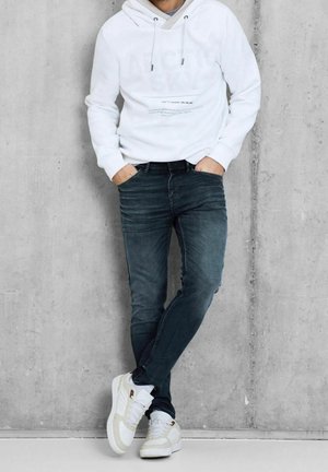 Man die een witte hoodie draagt, donkerblauwe slim-fit jeans, witte sneakers, staand met gekruiste benen en handen in de zakken tegen een betonnen muur.