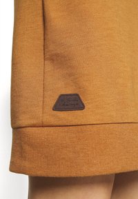 Orange sweatshirt fabric with a ribbed hem, featuring a brown rectangular brand label on the lower left side.