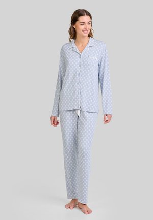 Mujer de pie descalza, vistiendo una camisa de pijama de manga larga abotonada de color azul claro con un patrón y pantalones a juego, sonriendo y mirando hacia un lado.