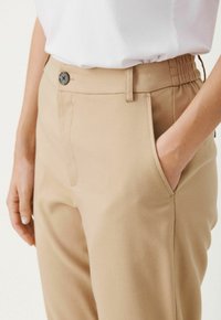 Beige skräddarsydda byxor med en mjuk textur, som har framknapp, sidofickor och ett elastiskt midjeband för extra komfort.