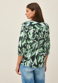 Blouse florale en vert foncé avec de grands motifs de feuilles en vert clair. Présente des manches trois-quarts avec des poignets froncés. Coupe décontractée.